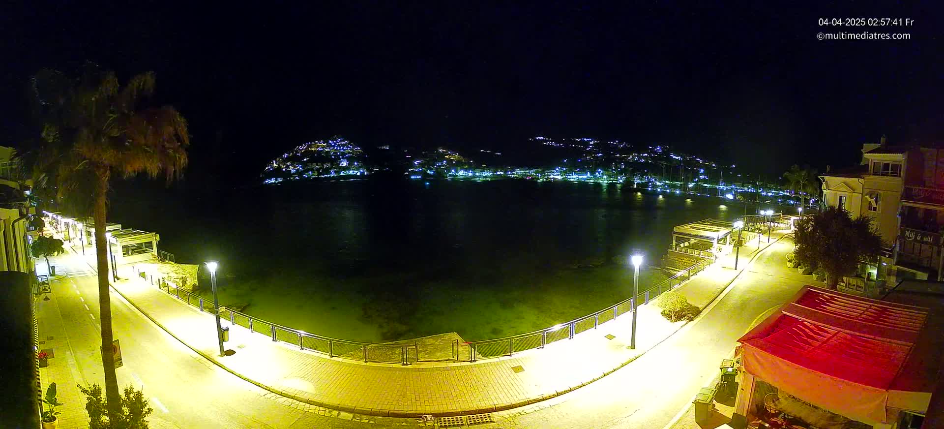 A nighttime, illuminated waterfront scene shows a calm body of water with a town's lights visible on the hills in the background.