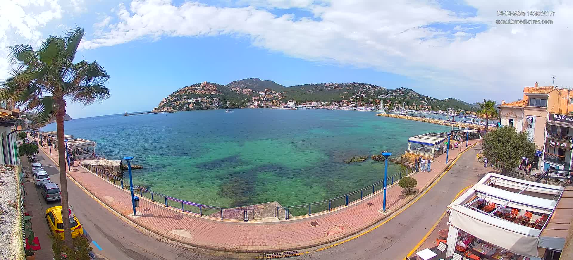 A partly cloudy day shows a seaside town with clear, teal water, a paved walkway, parked cars, and buildings along the coast, with a hill covered in buildings in the background.