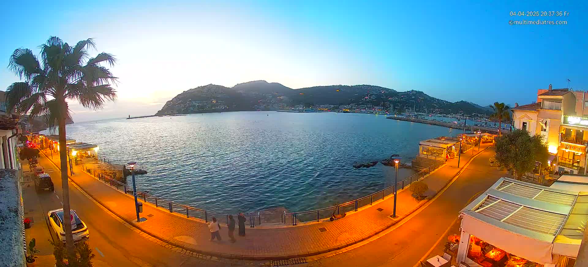 A calm, twilight-lit harbor scene shows a crescent-shaped waterfront walkway lined with restaurants and palm trees, overlooking a body of water and a hilltop town under a clear sky.
