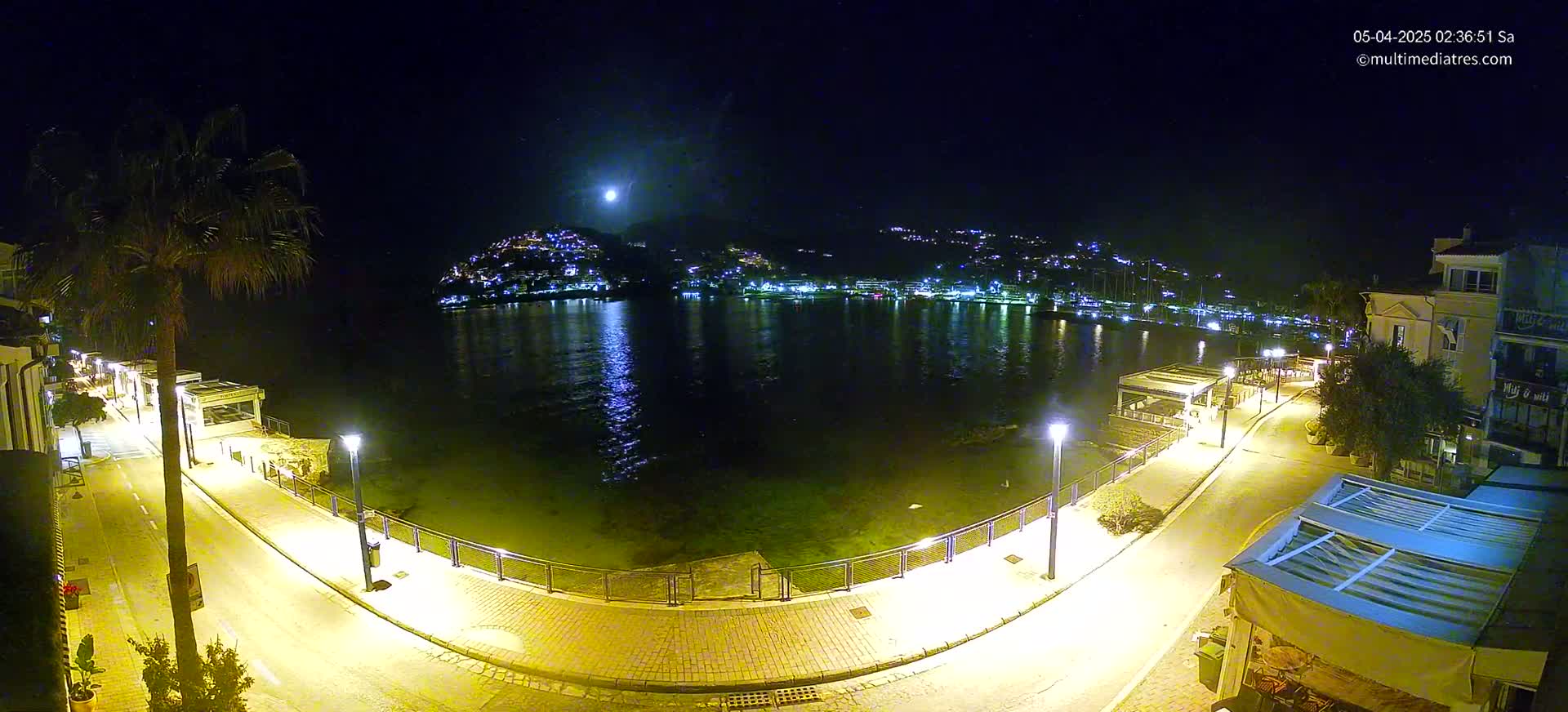 A nighttime, moonlit view of a calm harbor with a town lit up on the hills across the water, seen from a paved waterfront path lined with lights and buildings.