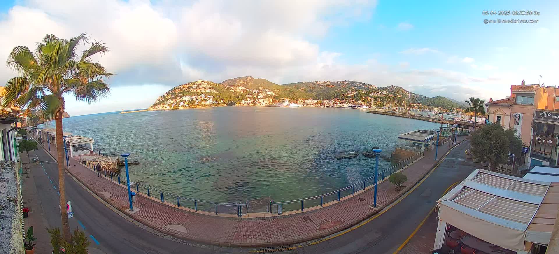 A partly cloudy day overlooks a calm bay with a town and hills in the background, seen from a waterfront promenade.