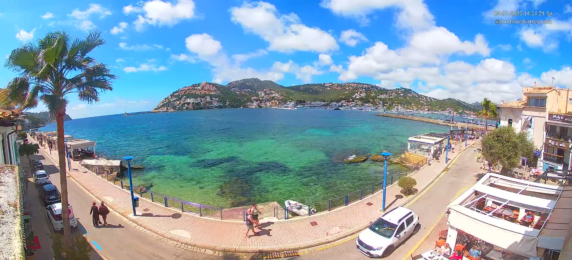 A sunny day overlooking a clear, turquoise bay with a waterfront promenade, buildings, and parked cars, under a partly cloudy sky.