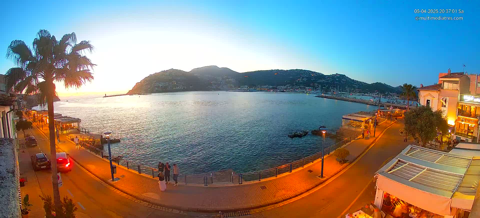 A waterfront scene at sunset shows a calm bay with boats, a paved walkway lined with restaurants and streetlights, and a hill overlooking the harbor.