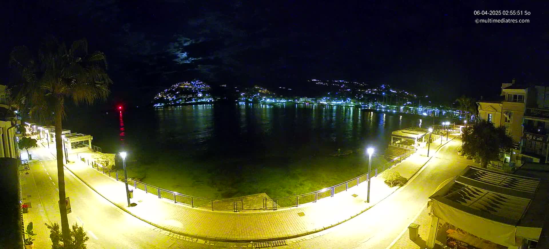 A nighttime view of a calm, illuminated waterfront with a paved walkway, buildings, and a hill in the distance under a partly cloudy moonlit sky.