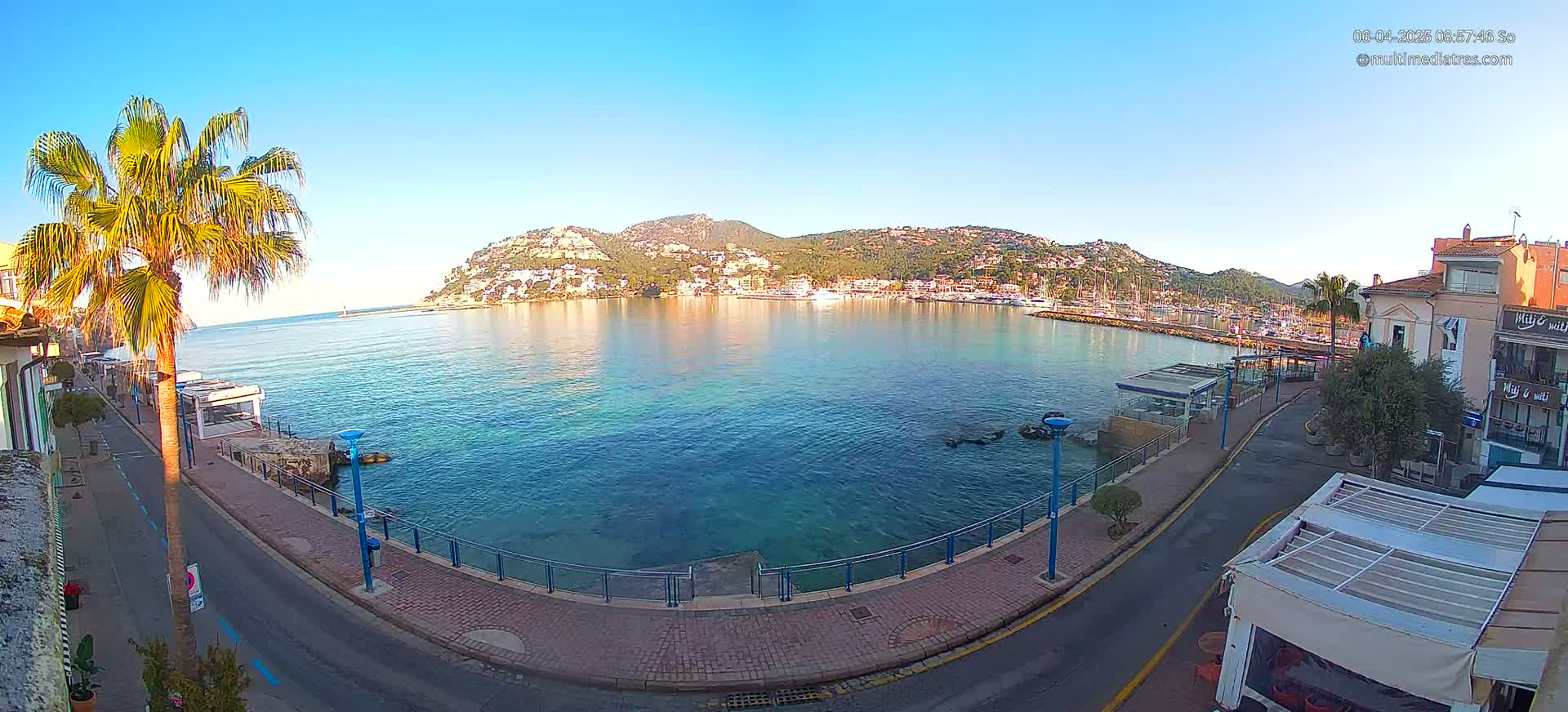 A calm, clear day reveals a harbor filled with boats, backed by a hillside town nestled against a mountain range, all viewed from a seaside promenade.