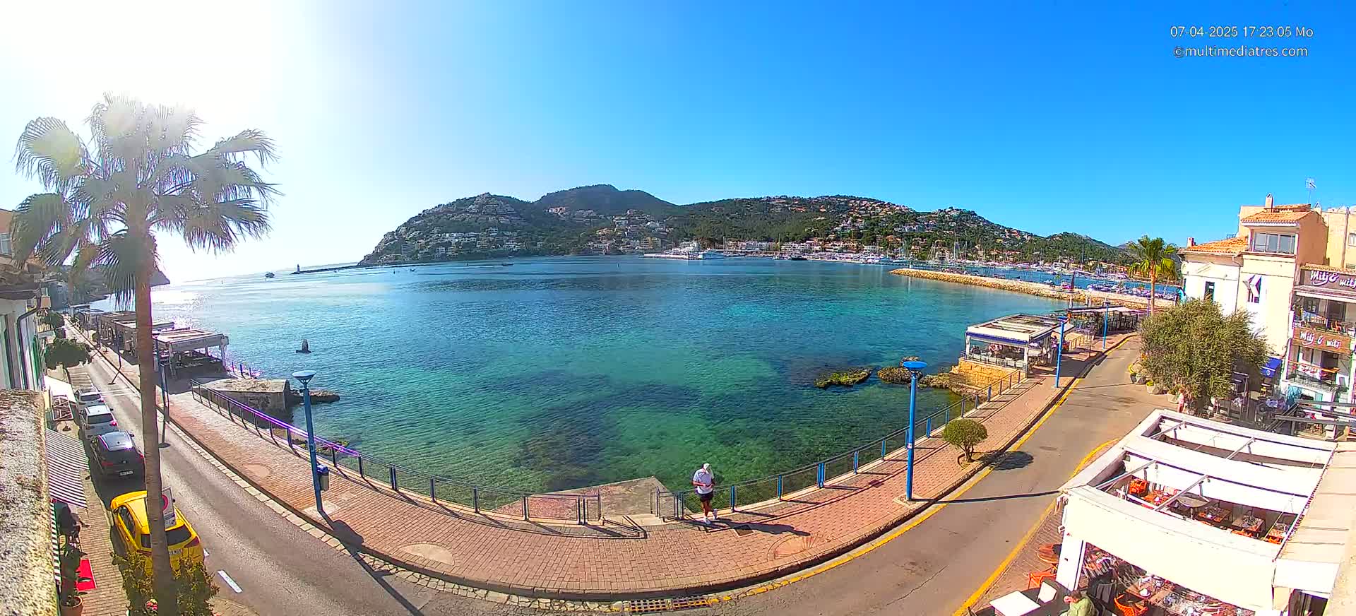A sunny day shows a clear turquoise harbor with a town and hill in the background, and a person walking along a paved walkway next to the water.