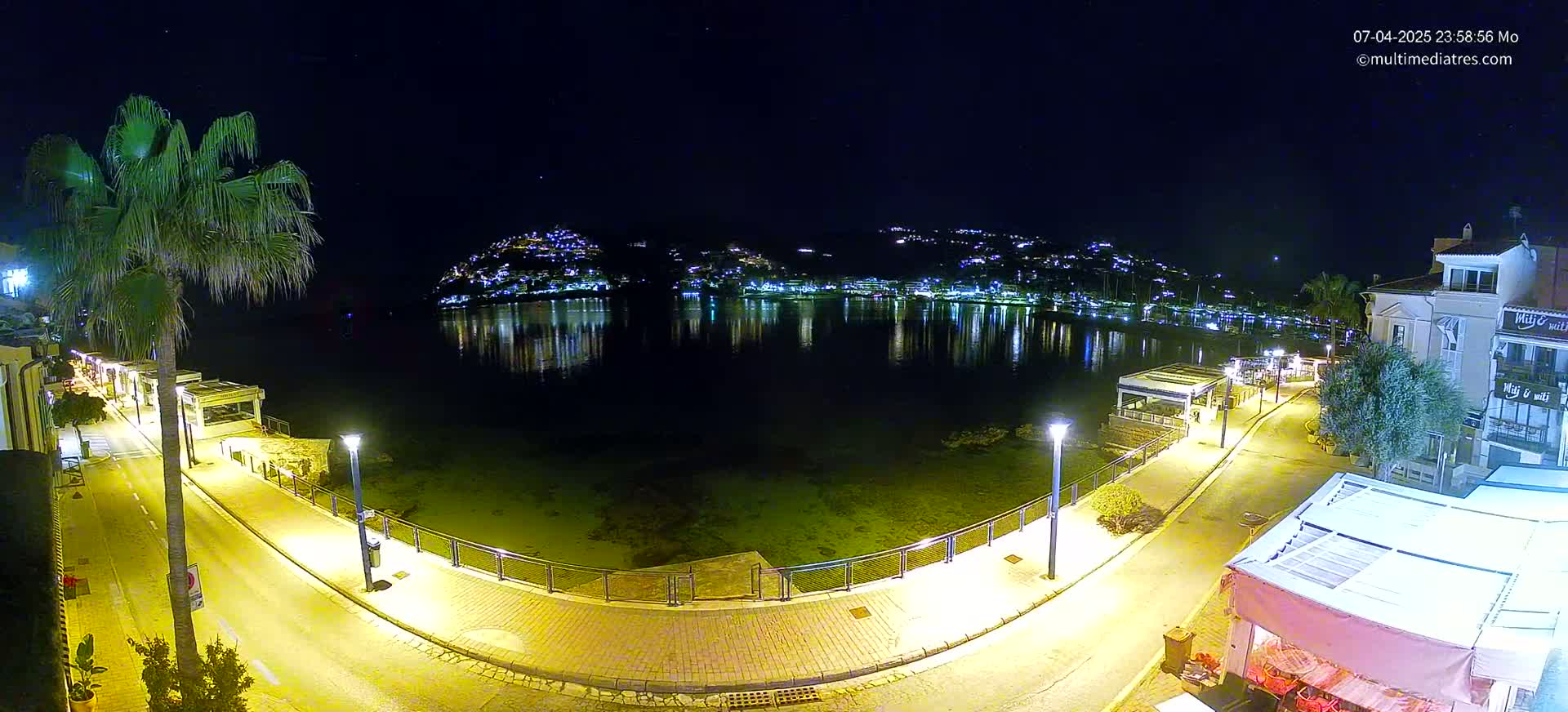 A nighttime view of a calm bay reflecting the lights of a town nestled on a hillside, with a paved walkway and palm trees lining the waterfront.