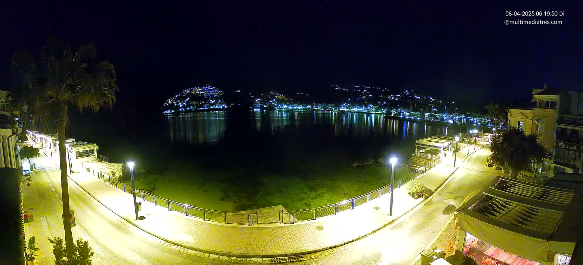 A nighttime, illuminated waterfront scene shows a calm body of water reflecting lights from buildings on a hill across the water, with a paved walkway and palm trees lining the shore.