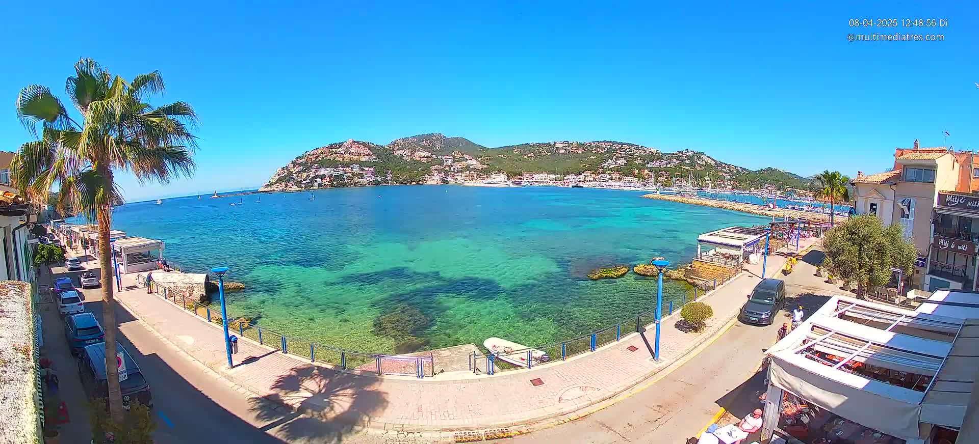 A sunny day reveals a clear turquoise bay with a hillside town and a palm tree-lined waterfront promenade.