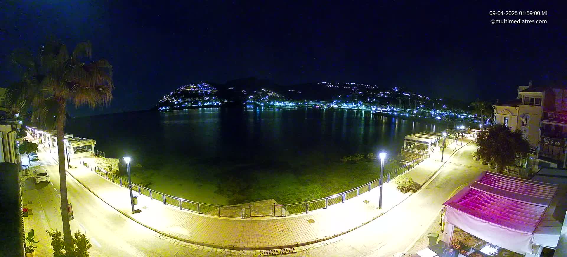 A nighttime, seaside town scene shows a calm body of water reflecting the lights of buildings on a hill across the water, with a paved walkway and palm trees illuminated along the shoreline.