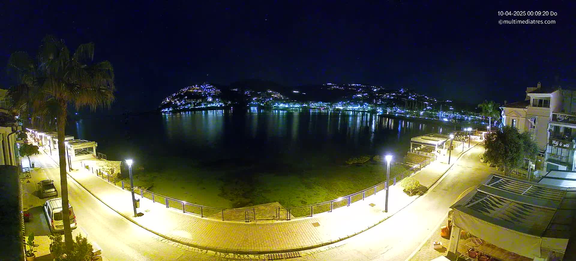 A nighttime view of a calm bay, illuminated by city lights on the opposite shore, with a paved walkway and buildings lining the waterfront under a clear, starry sky.