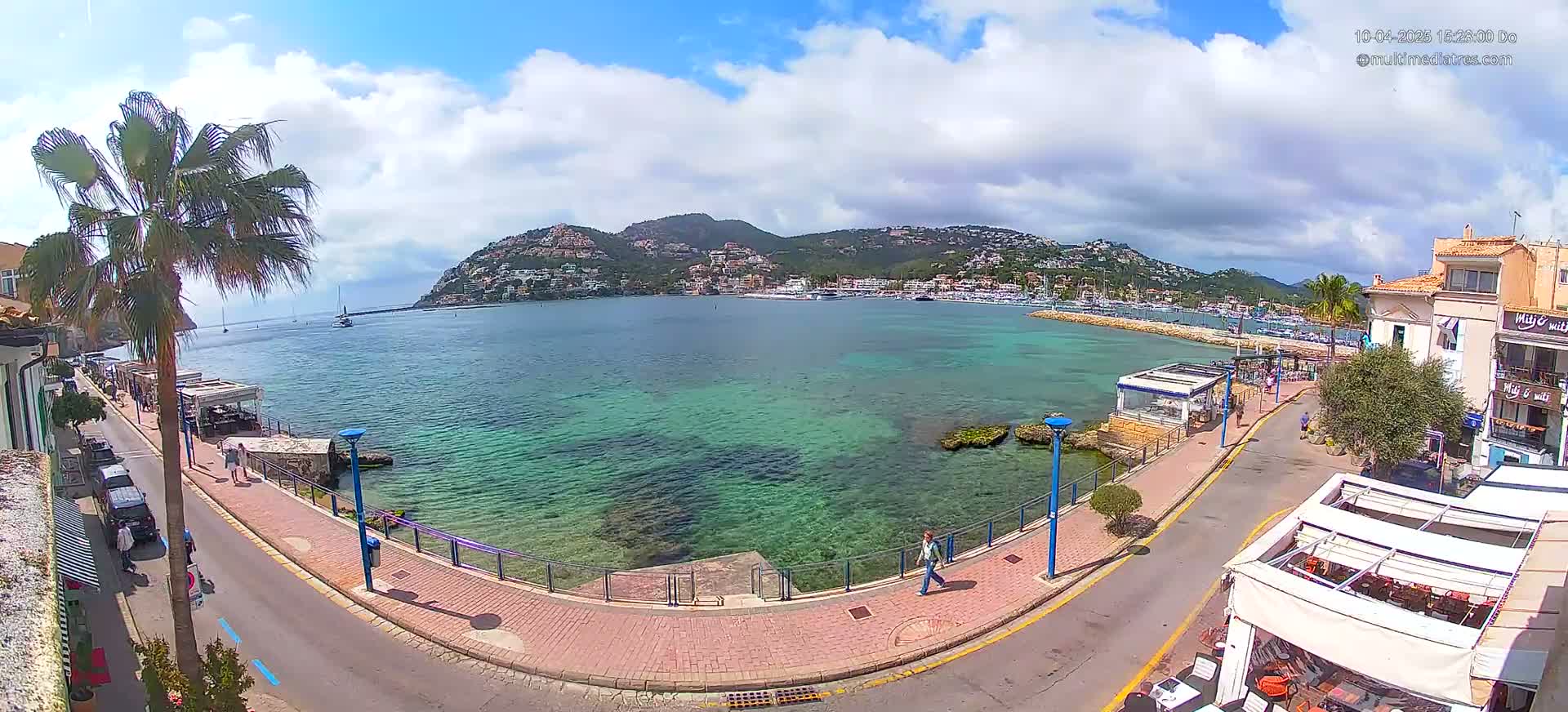 A partly cloudy day overlooks a calm, clear, teal-colored harbor with buildings and a few people walking along a seaside promenade.