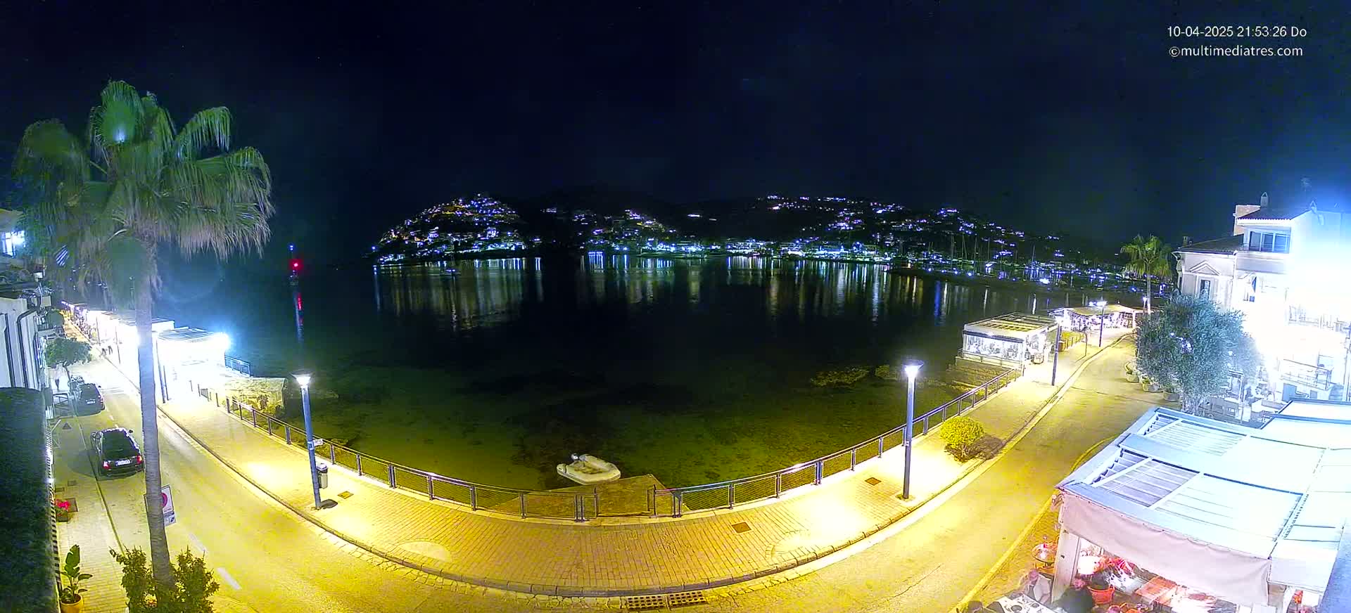 A nighttime view of a calm, dark-water harbor reflecting the lights of a town on a hillside, with a paved walkway and palm trees lining the waterfront.