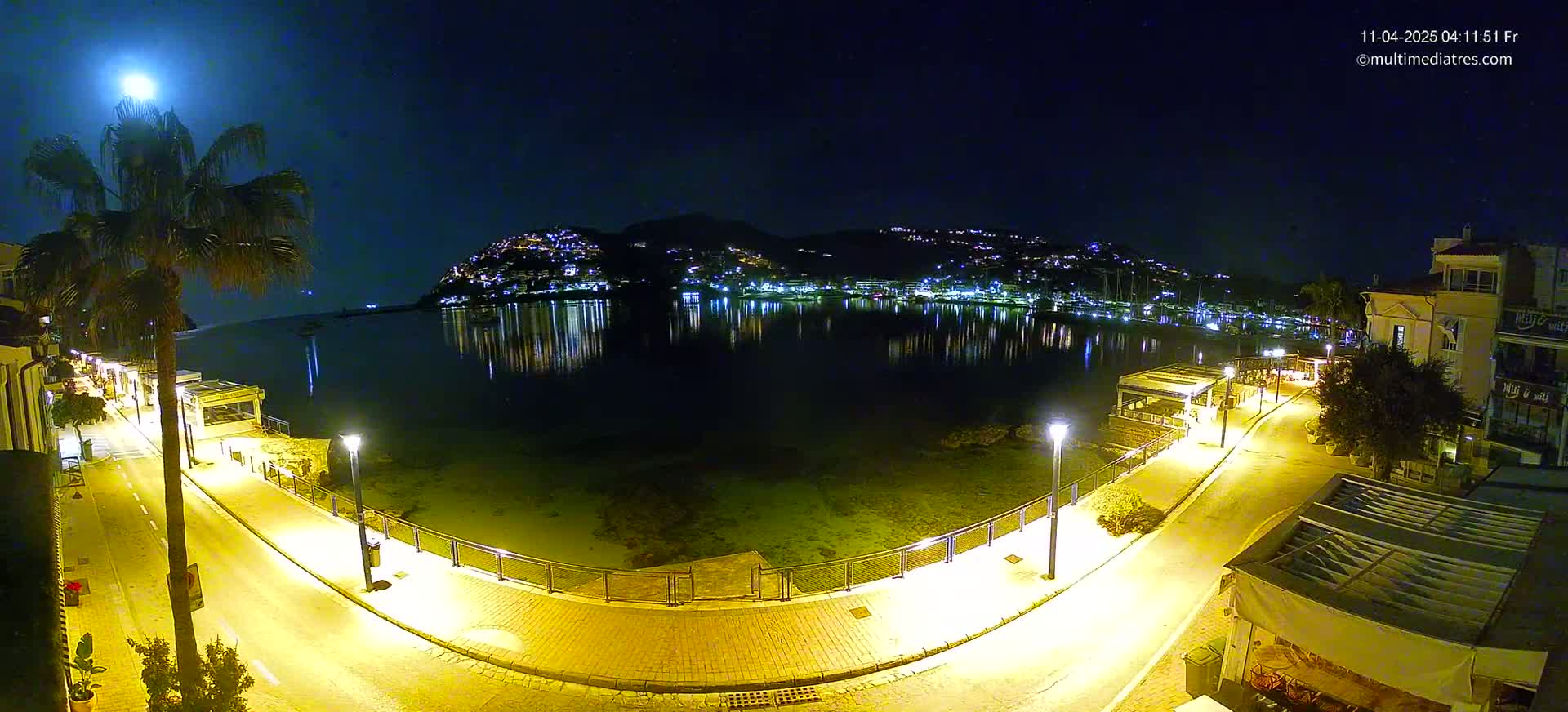 A moonlit night scene shows a waterfront promenade illuminated by streetlights, with a calm body of water reflecting the lights of buildings on a hillside across the water.