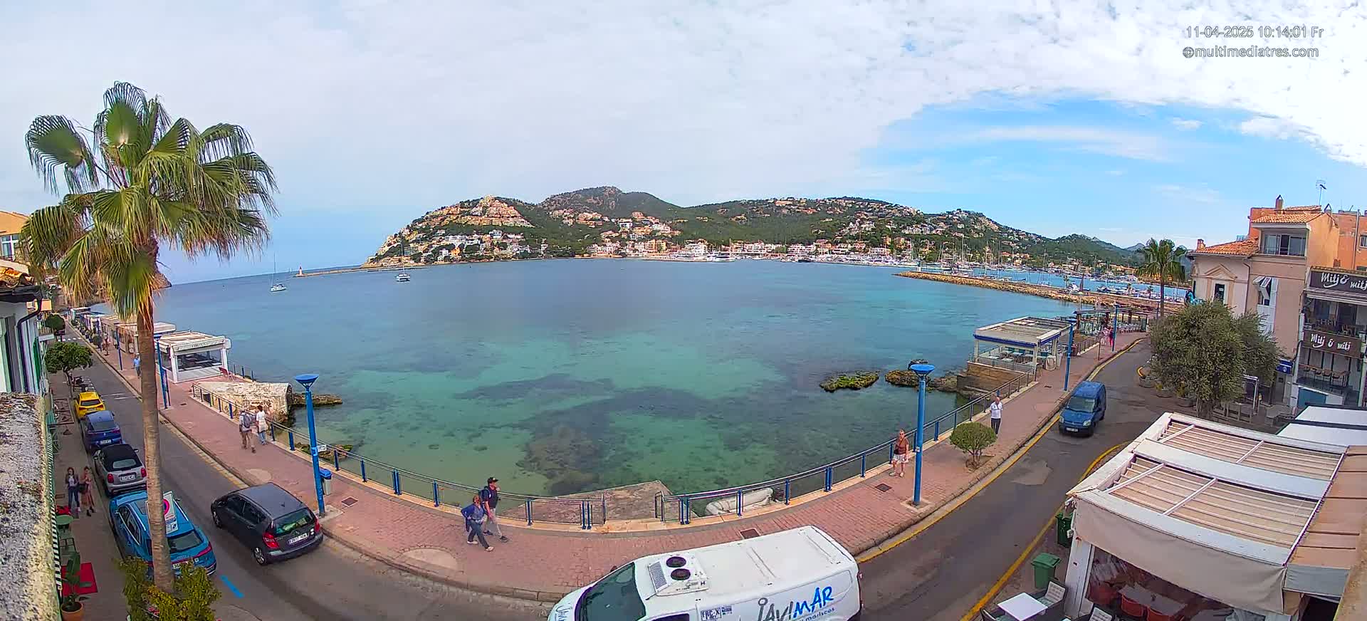 A seaside town with clear turquoise water, a waterfront promenade, parked cars, and buildings sits under a partly cloudy sky.