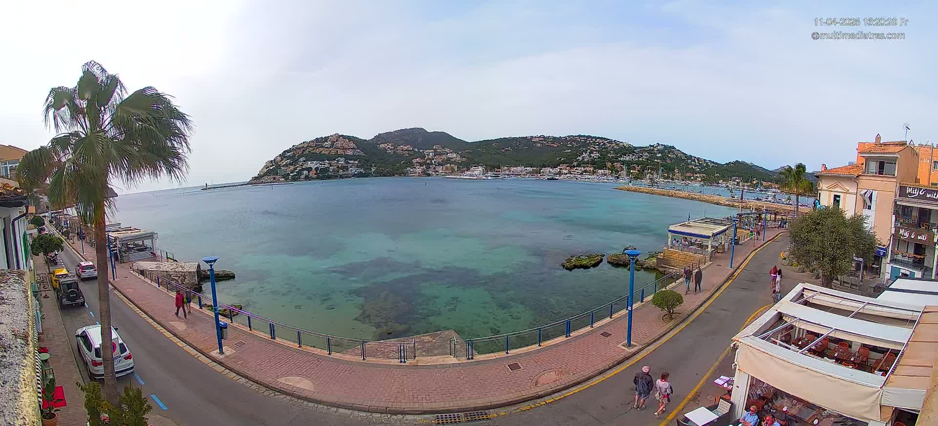 A calm, clear day overlooks a harbor town with a hill covered in buildings in the background, cars parked along a waterfront road, and several people walking along a promenade.