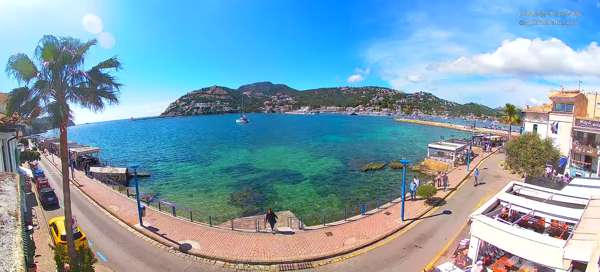 A sunny day reveals a seaside town with clear turquoise water, a paved walkway, buildings, and palm trees, all set against the backdrop of a hilly coastline.