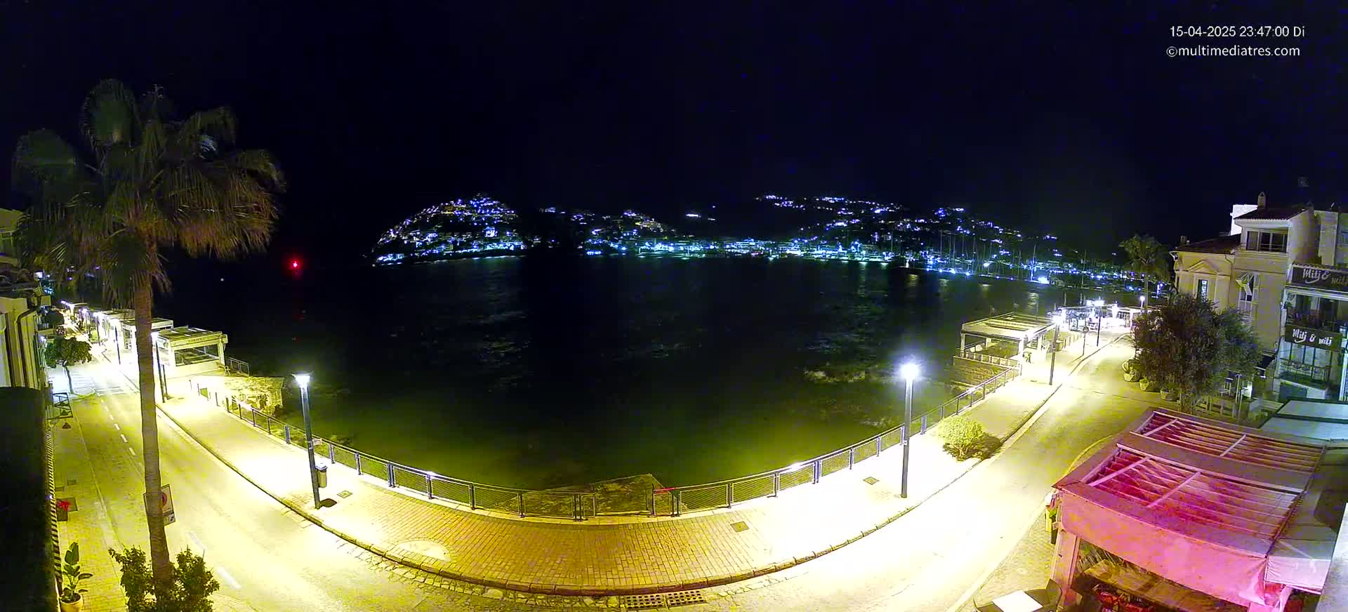A nighttime view of a waterfront promenade, illuminated by streetlights, overlooking a calm bay with a hilltop town glowing in the distance.