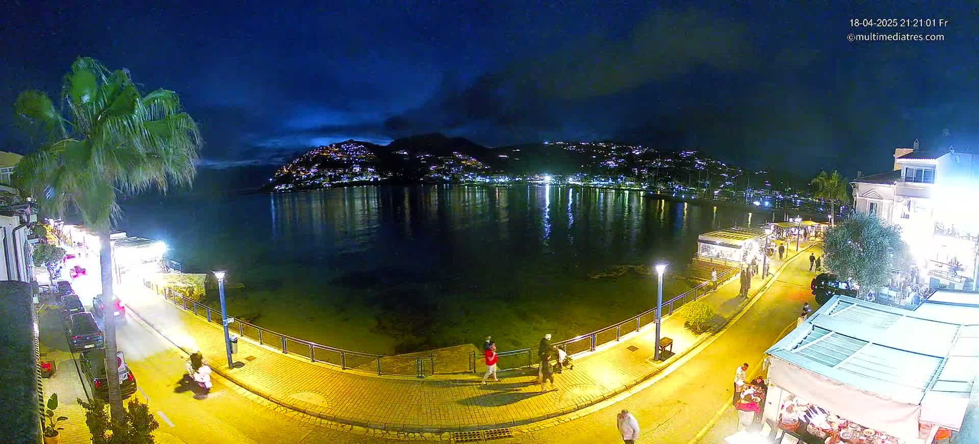 A nighttime seaside town with a calm, dark-blue body of water reflecting the lights of buildings on a hill, a paved walkway with people strolling, and palm trees lining the street.