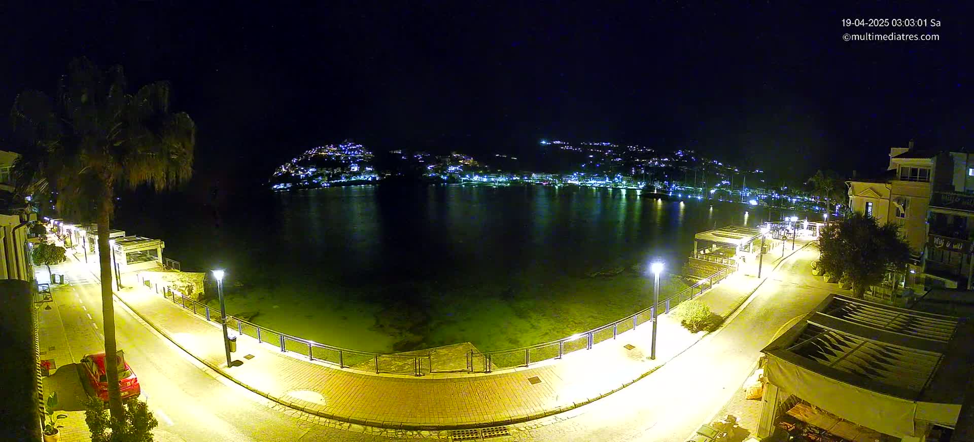 A nighttime, illuminated waterfront scene shows a calm body of water reflecting a hilltop town across the bay, with a paved walkway and buildings lining the shoreline.