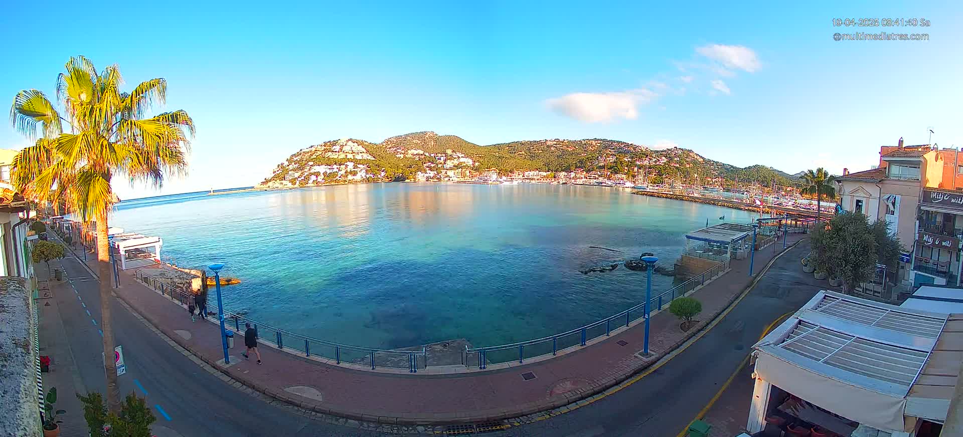 A clear, sunny day shows a seaside town with a calm, turquoise bay, a palm tree-lined promenade, and hills covered in buildings and vegetation in the background.