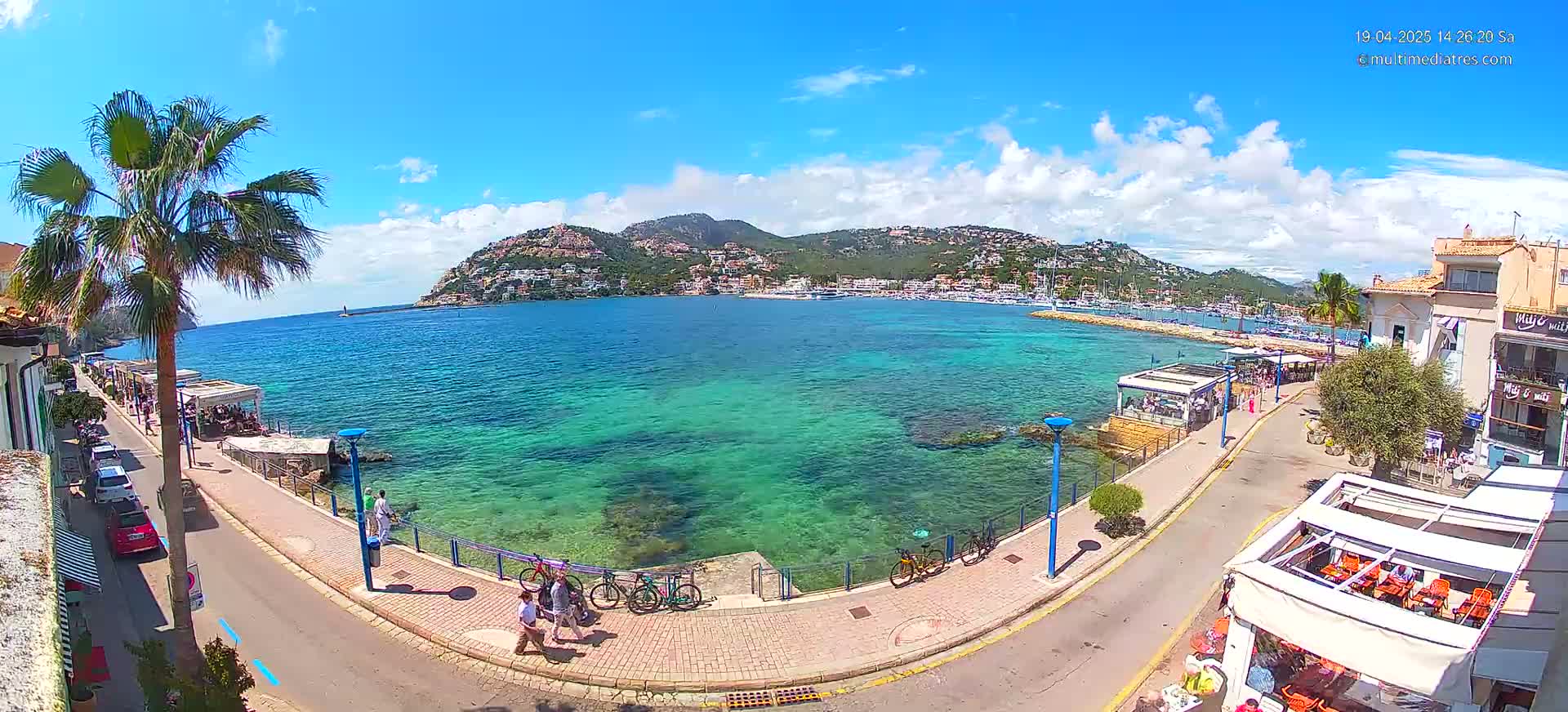 A sunny day reveals a coastal town with a clear, teal-colored bay, bordered by a paved walkway lined with buildings and palm trees, and hills in the background.