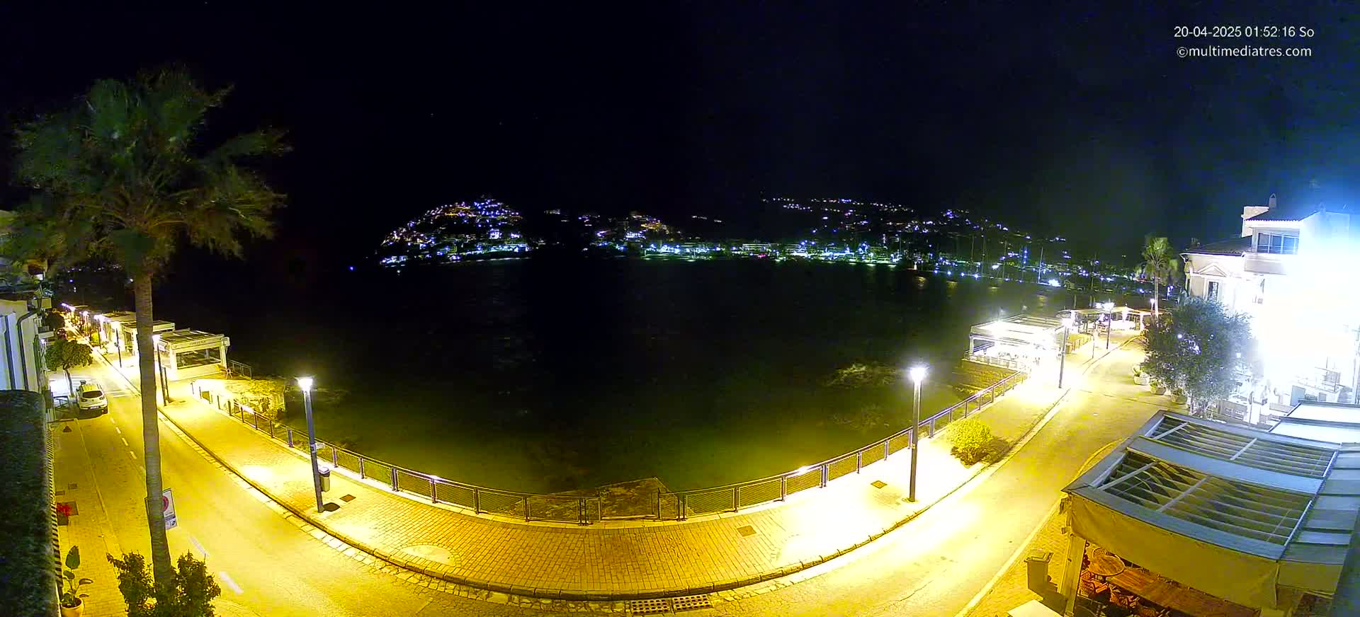 A nighttime view of a waterfront area with a calm body of water, illuminated streets, and buildings along the shore under a clear, dark sky.