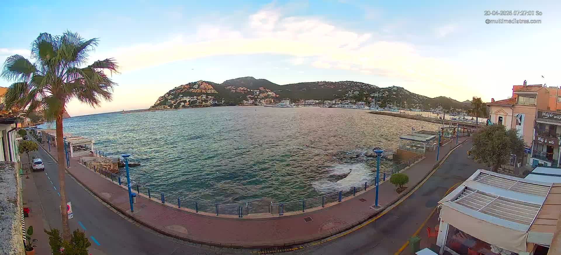A calm, clear morning scene shows a waterfront road curving past a harbor filled with boats, under a partly cloudy sky, with a hill covered in buildings in the background.