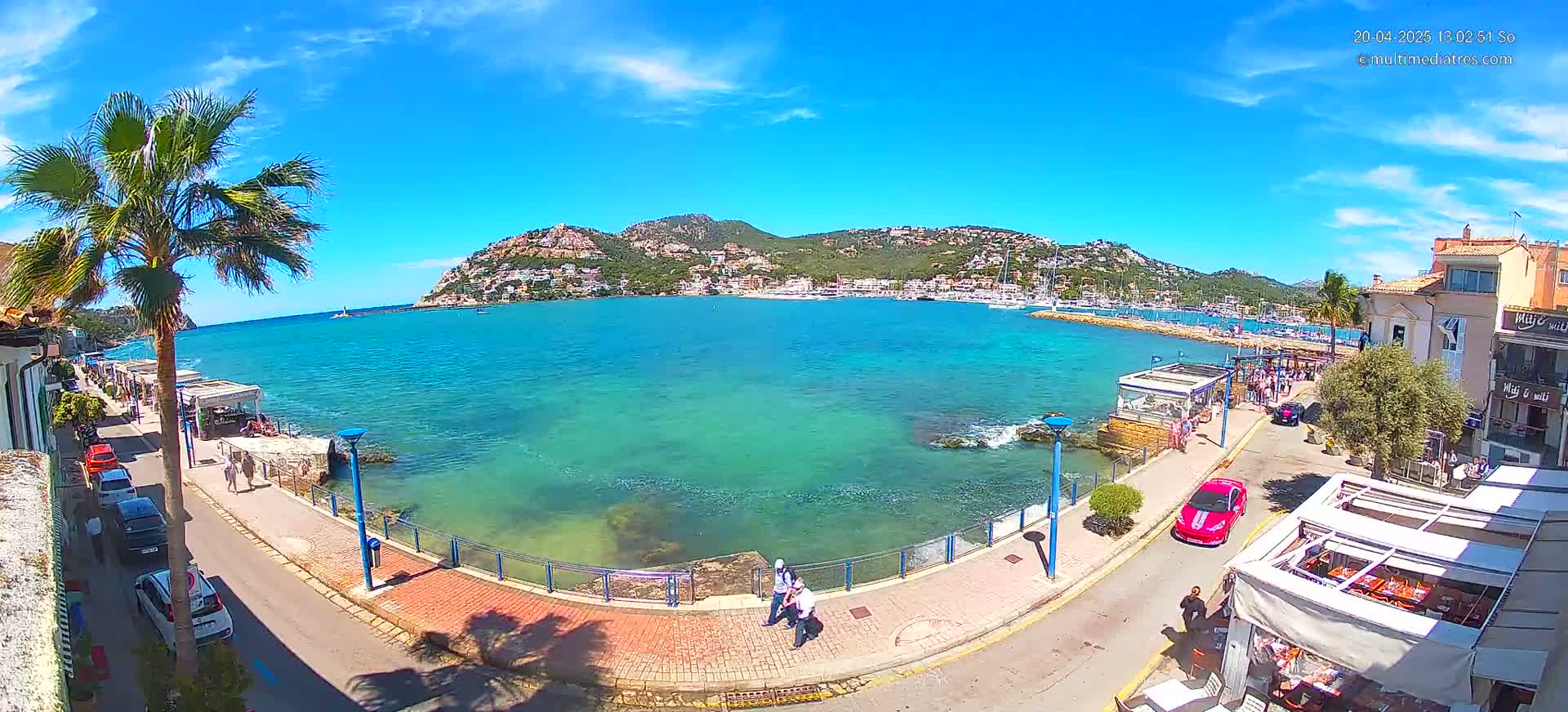 A sunny day shows a coastal town's waterfront with turquoise water, a marina filled with boats, and hills in the background.