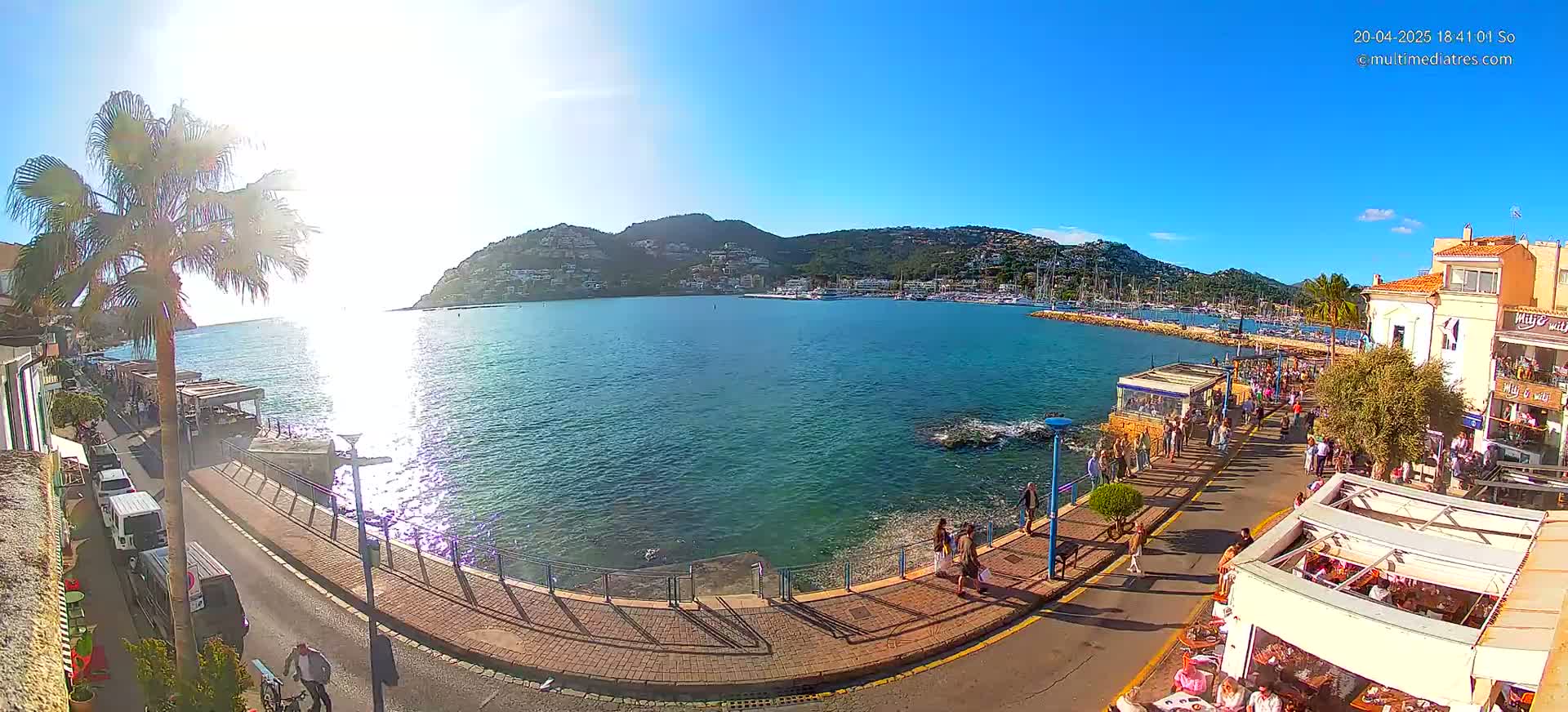 A sunny day overlooking a harbor filled with boats, a waterfront promenade with people walking, and a hillside town in the background.