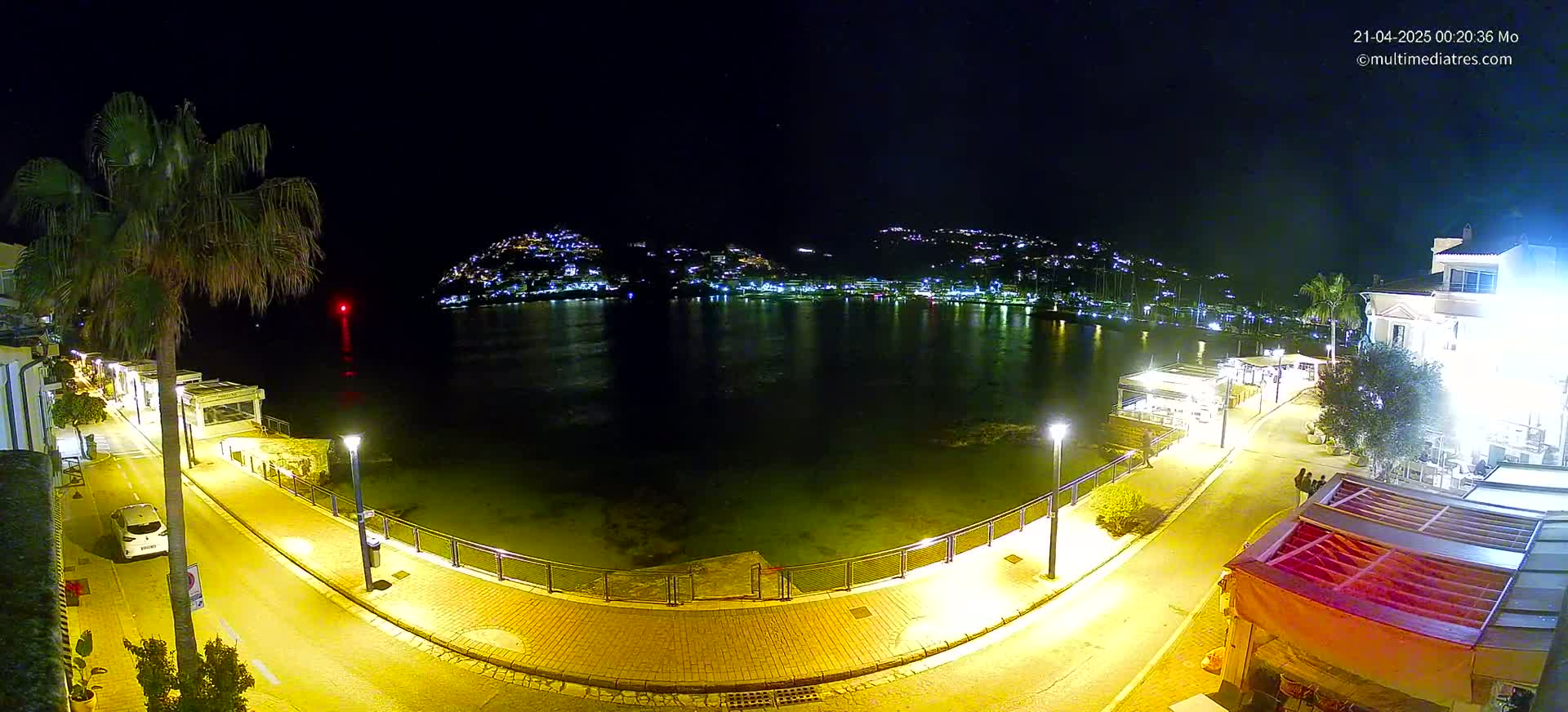 A nighttime, illuminated waterfront scene shows a calm body of water reflecting lights from buildings on the opposite shore and a paved walkway lined with restaurants and palm trees.