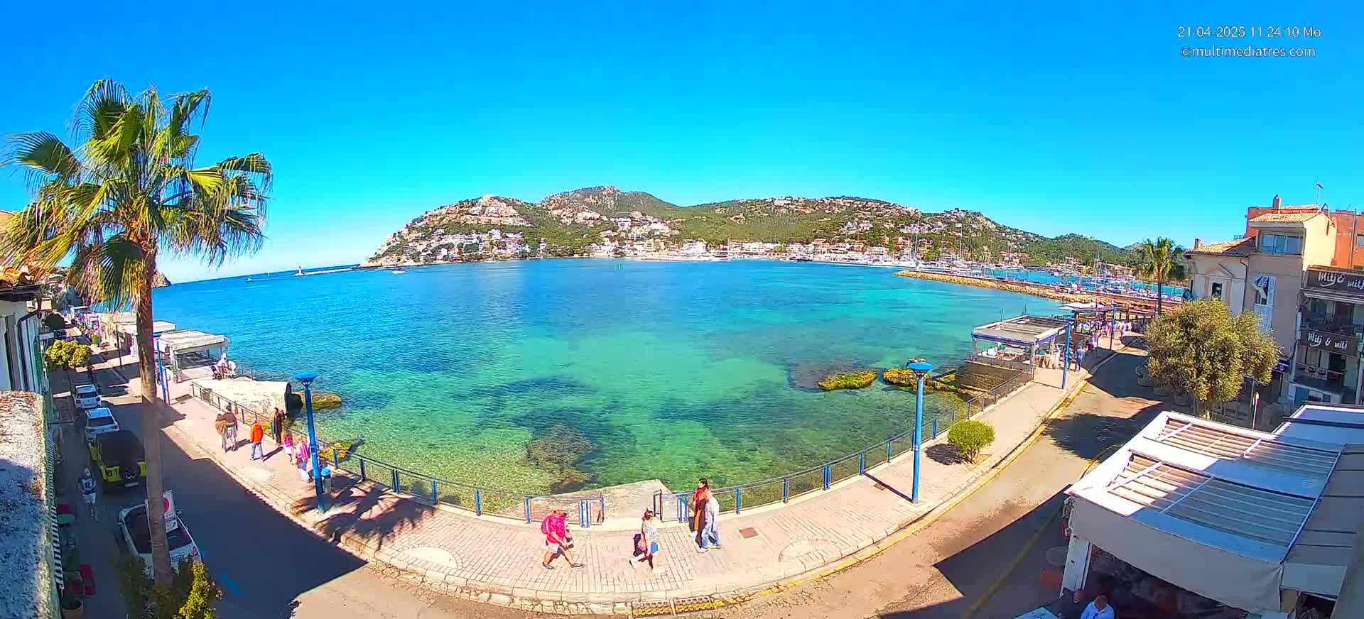 A clear, sunny day reveals a coastal town with turquoise water, a waterfront promenade, palm trees, and a few people walking along the shore, all nestled against a backdrop of green hills.