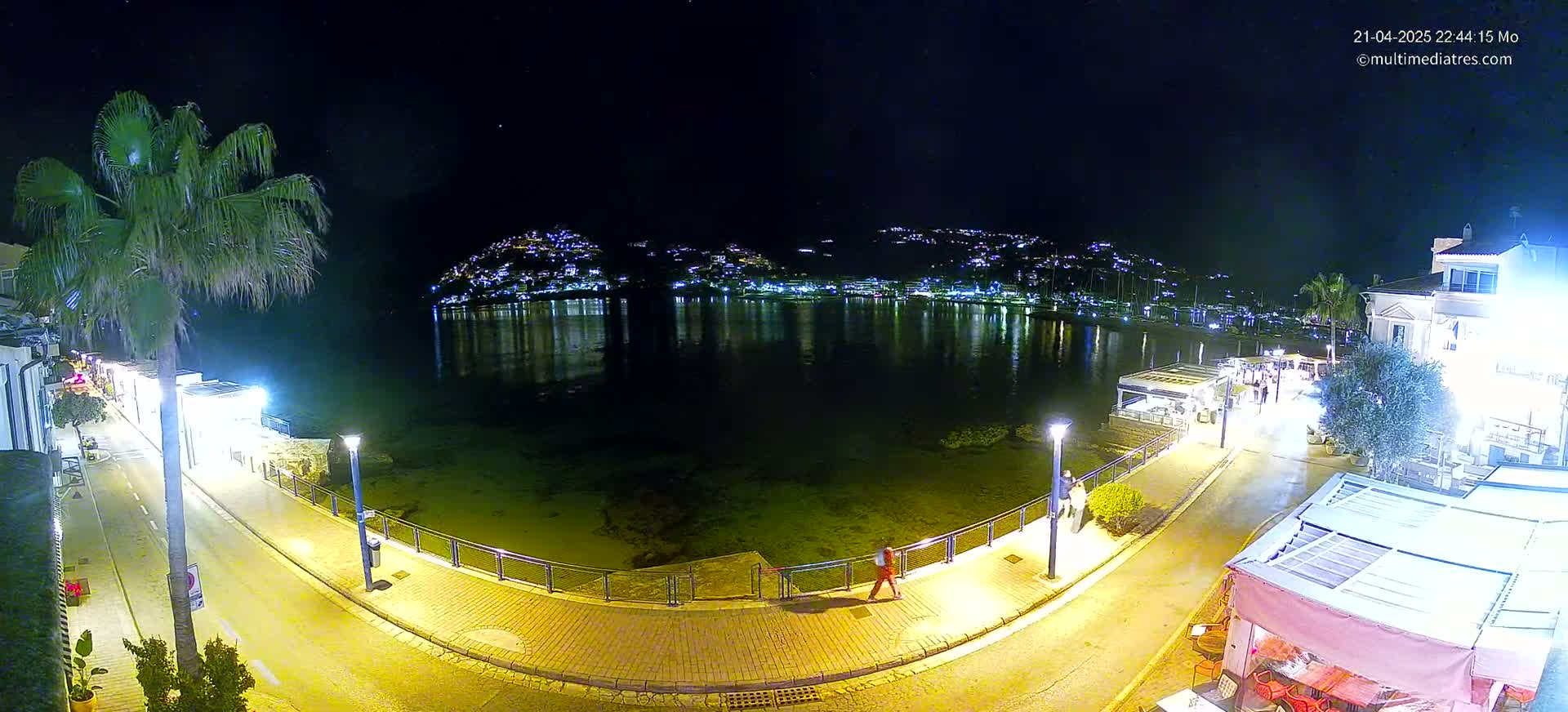 A nighttime view of a waterfront town, illuminated by streetlights, with a calm body of water reflecting the lights of buildings on a hill in the background.