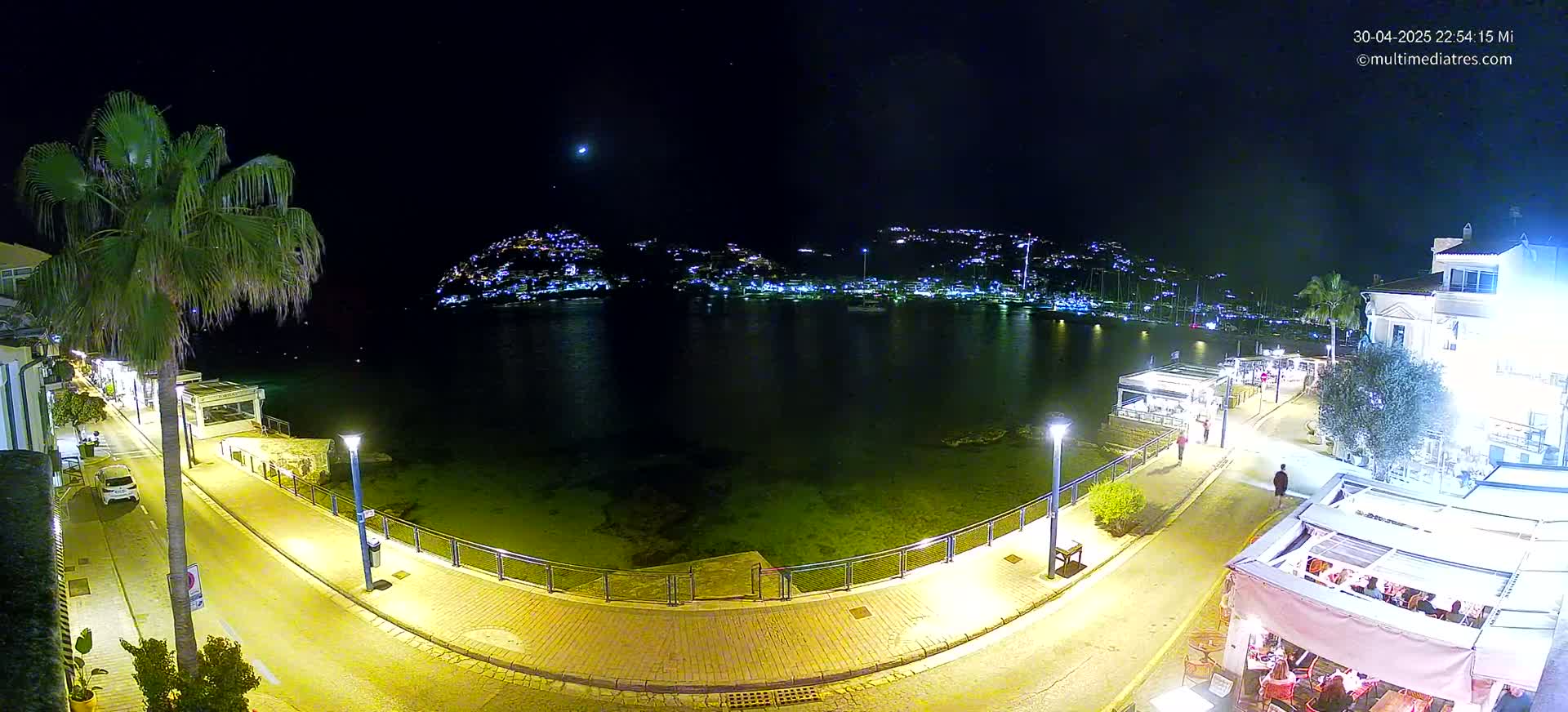 A nighttime view of a waterfront town with calm waters, illuminated buildings across the bay, and a paved walkway lined with palm trees and outdoor seating.