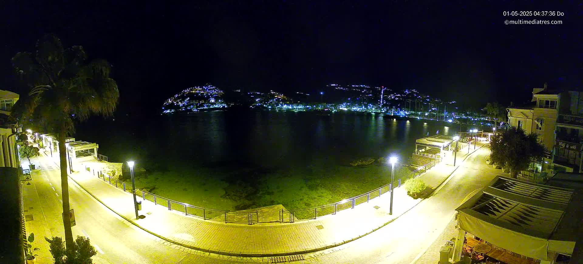 A nighttime, aerial view shows a calm bay with a town's lights across the water, a paved walkway curving along the waterfront illuminated by streetlights.