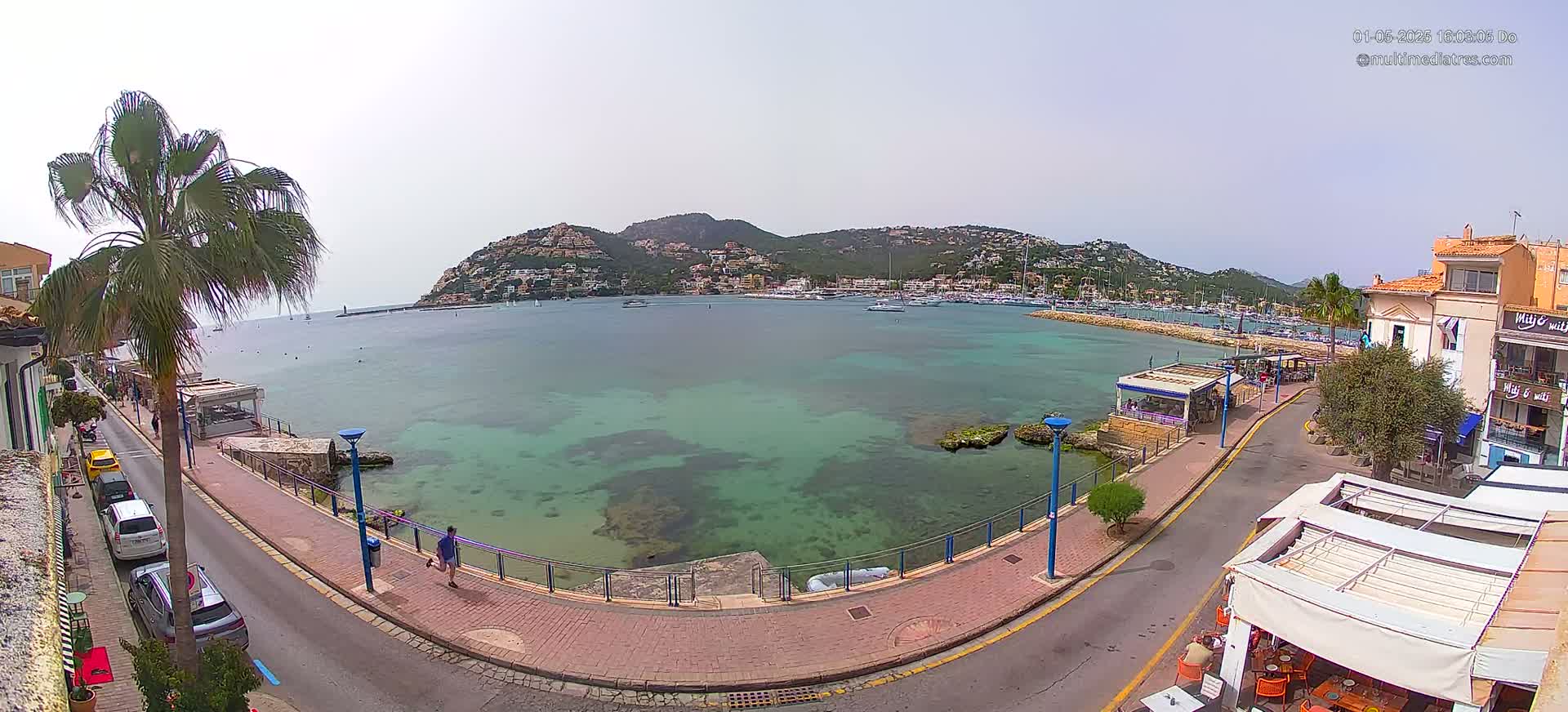 A calm, clear day reveals a harbor filled with boats nestled beneath a hillside town, seen from a high vantage point overlooking a paved waterfront walkway.