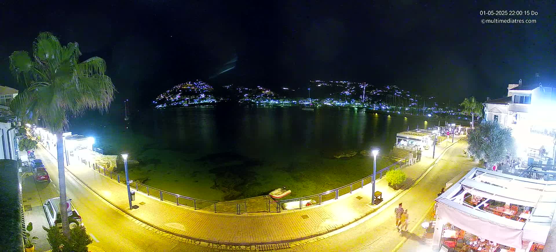 A nighttime view of a waterfront town with palm trees, illuminated buildings, a calm bay, and a few people walking along a paved walkway.