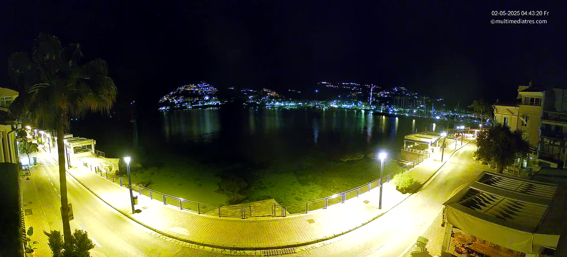 A nighttime view of a calm harbor, illuminated by streetlights, with a town nestled on a hill in the background.