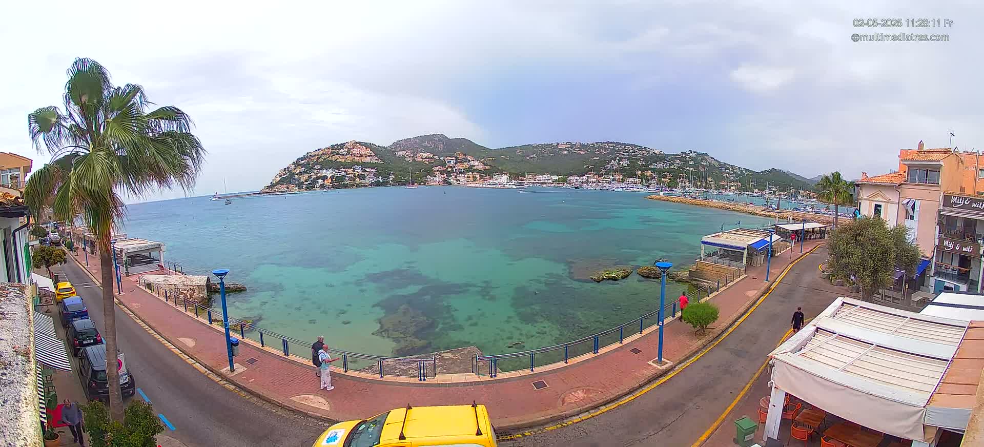A partly cloudy day reveals a waterfront scene with clear, teal water, a town nestled against a hillside, and a roadway lined with parked cars and small buildings.