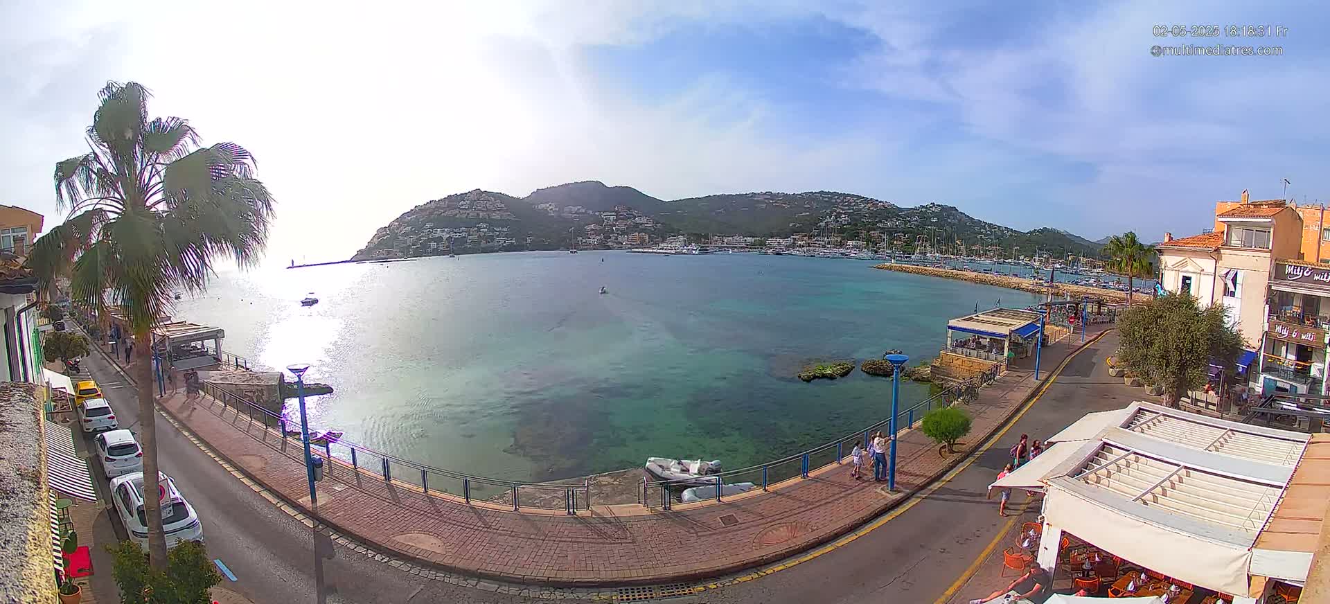 A sunny day shows a calm bay with boats in a harbor, surrounded by buildings and a paved walkway with people walking along the waterfront.