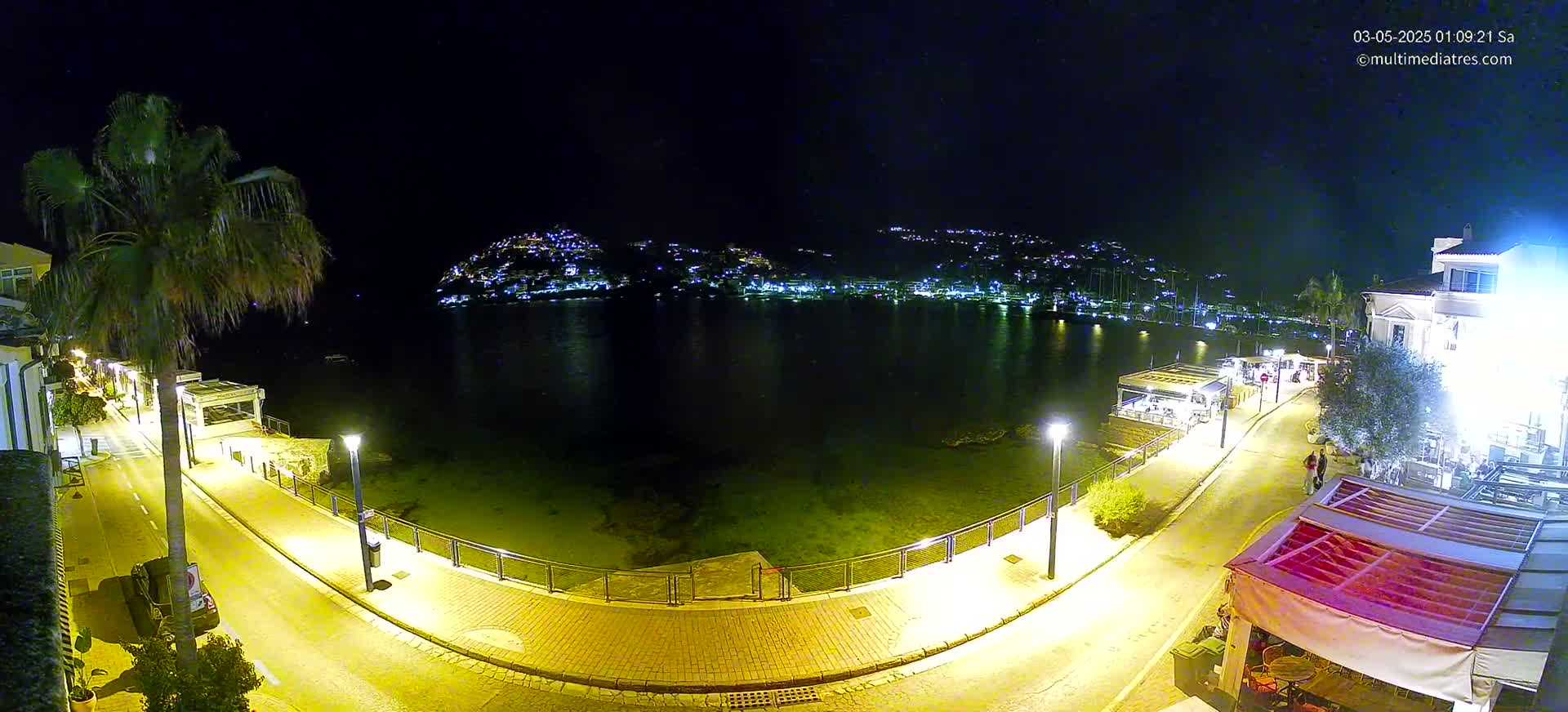 A nighttime, illuminated waterfront scene shows a calm body of water with a town's lights visible across it, a curved roadway with lamp posts lining the shore, and a palm tree near buildings.
