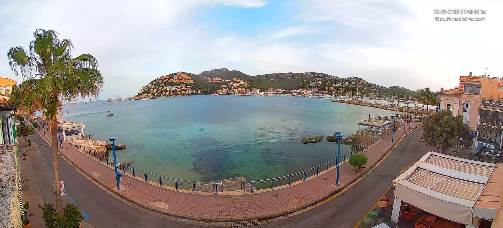 A partly cloudy day shows a calm bay with clear, shallow water, a paved waterfront promenade, and a hill with buildings in the background.
