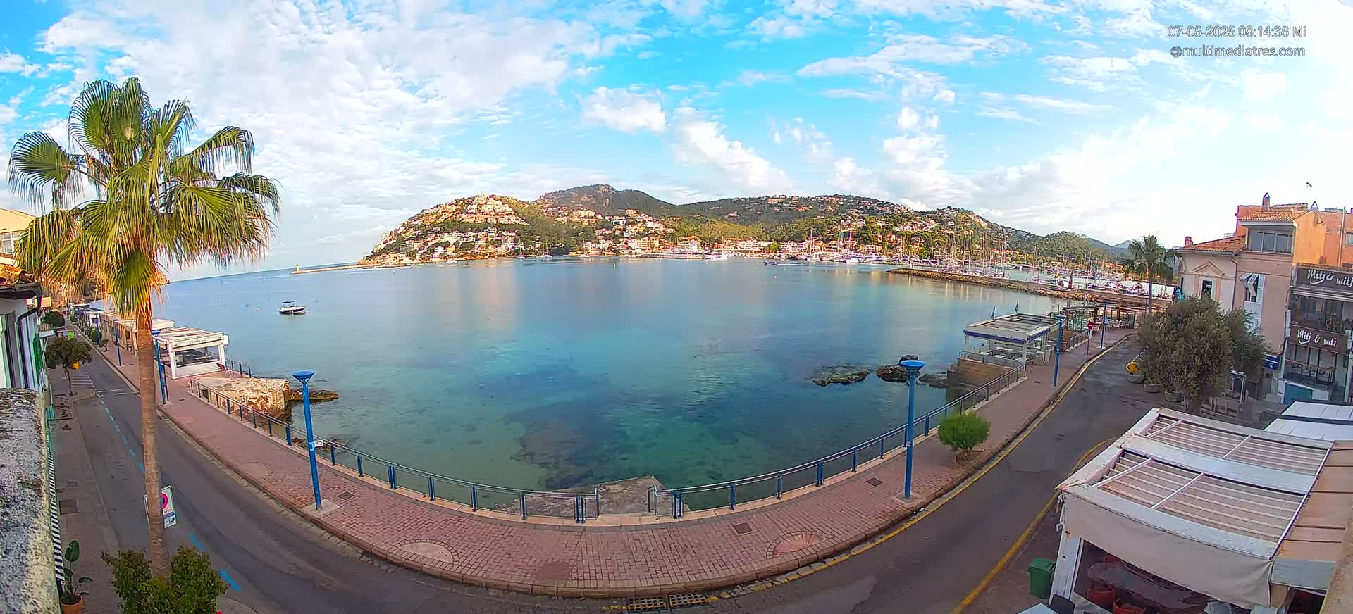 A partly sunny day shows a calm bay with clear turquoise water, a waterfront promenade with palm trees, and a hillside town in the background.