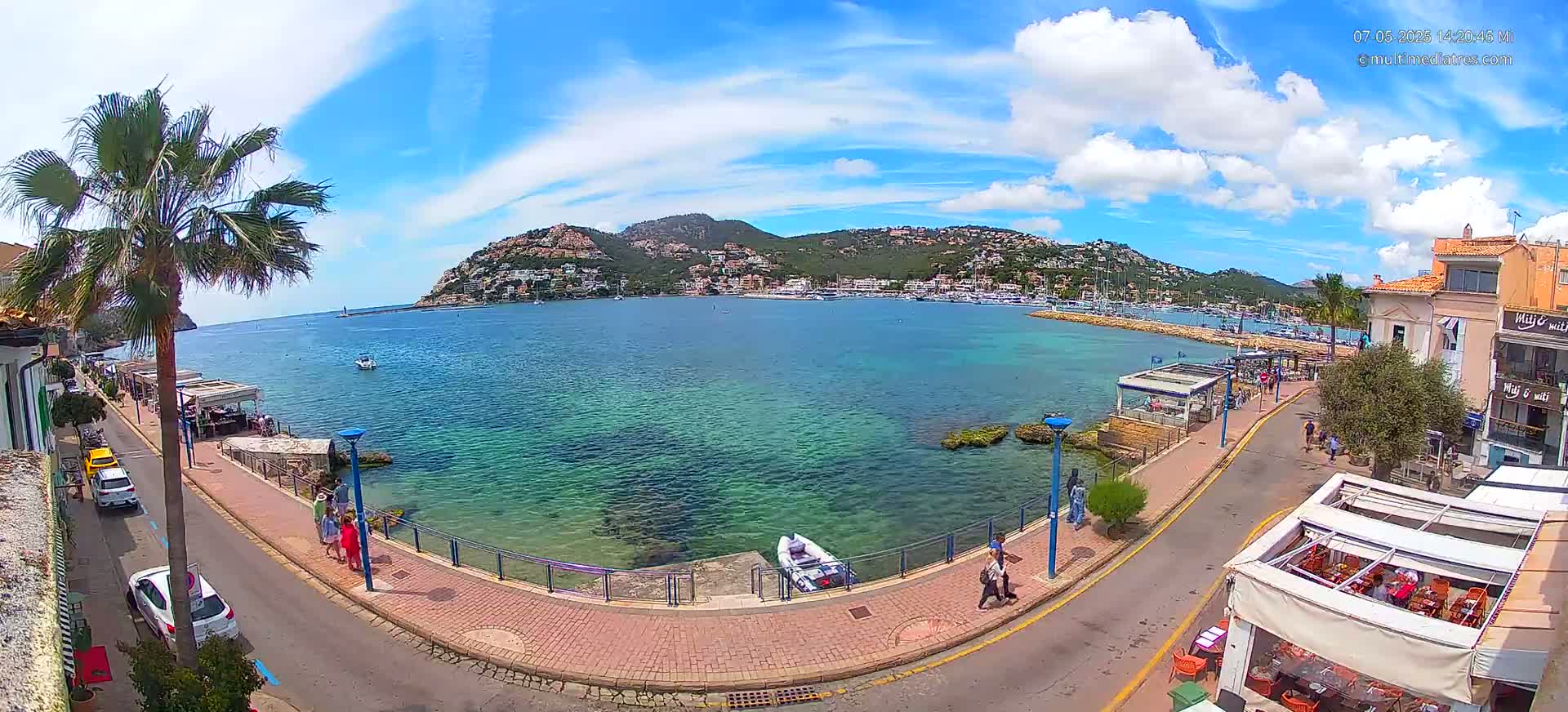 A sunny day overlooking a clear, turquoise harbor with a town nestled against a hillside, a palm tree in the foreground, and people strolling along a waterfront promenade.