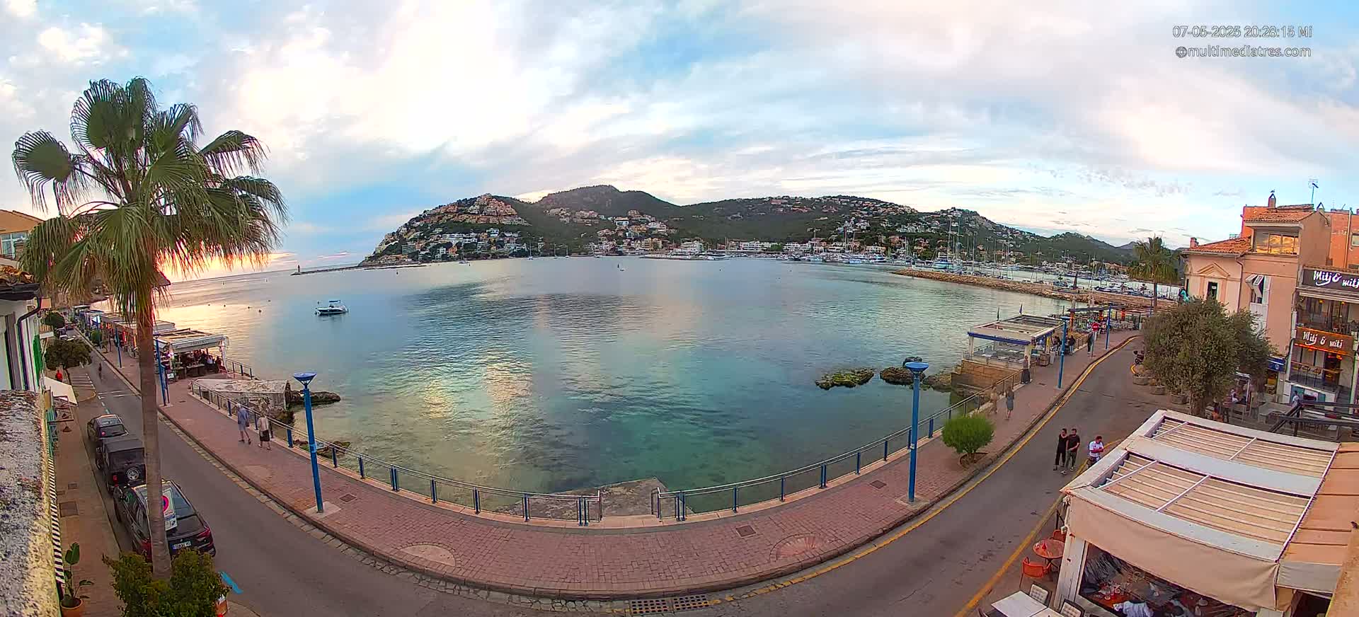 A calm bay with a hill covered in buildings is seen from a waterfront promenade at sunset under a partly cloudy sky.