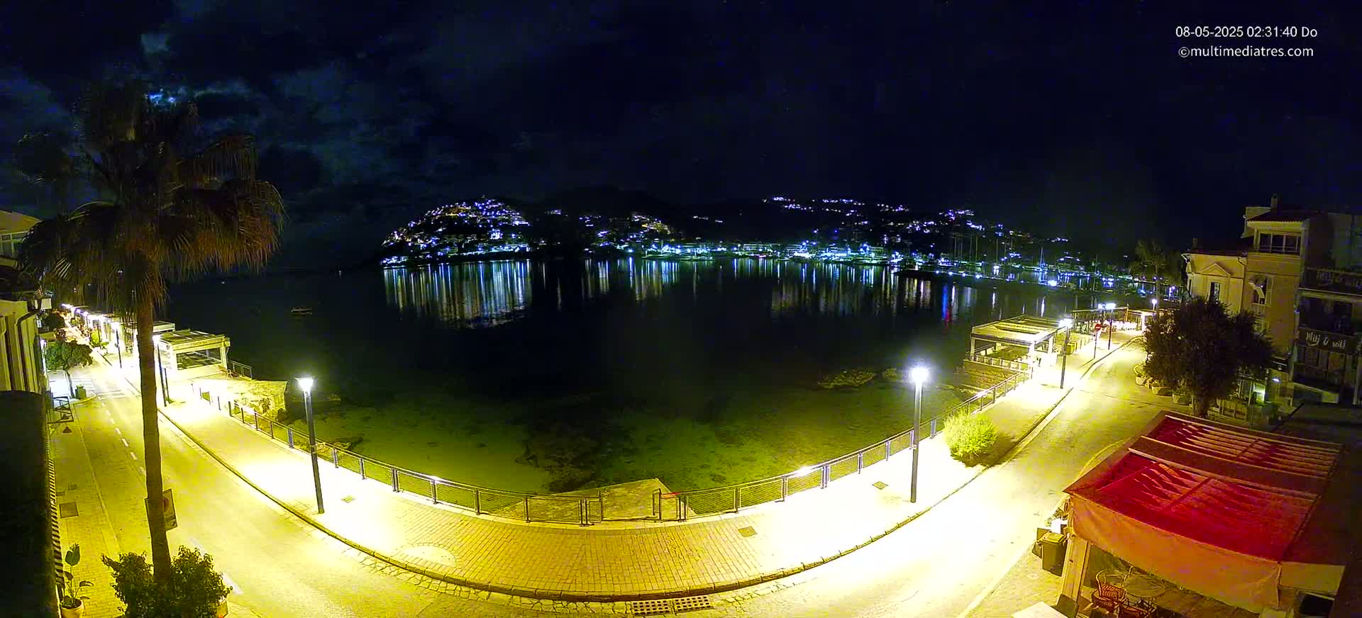 A nighttime view of a calm waterfront, illuminated by streetlights, with a hilltop town twinkling across the water under a partly cloudy sky.