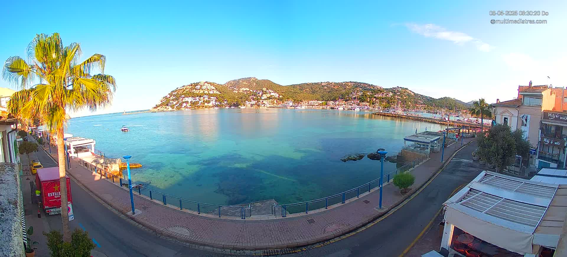 A clear, sunny day shows a calm bay with clear turquoise water, a town nestled against a hillside, and a palm tree beside a paved walkway.