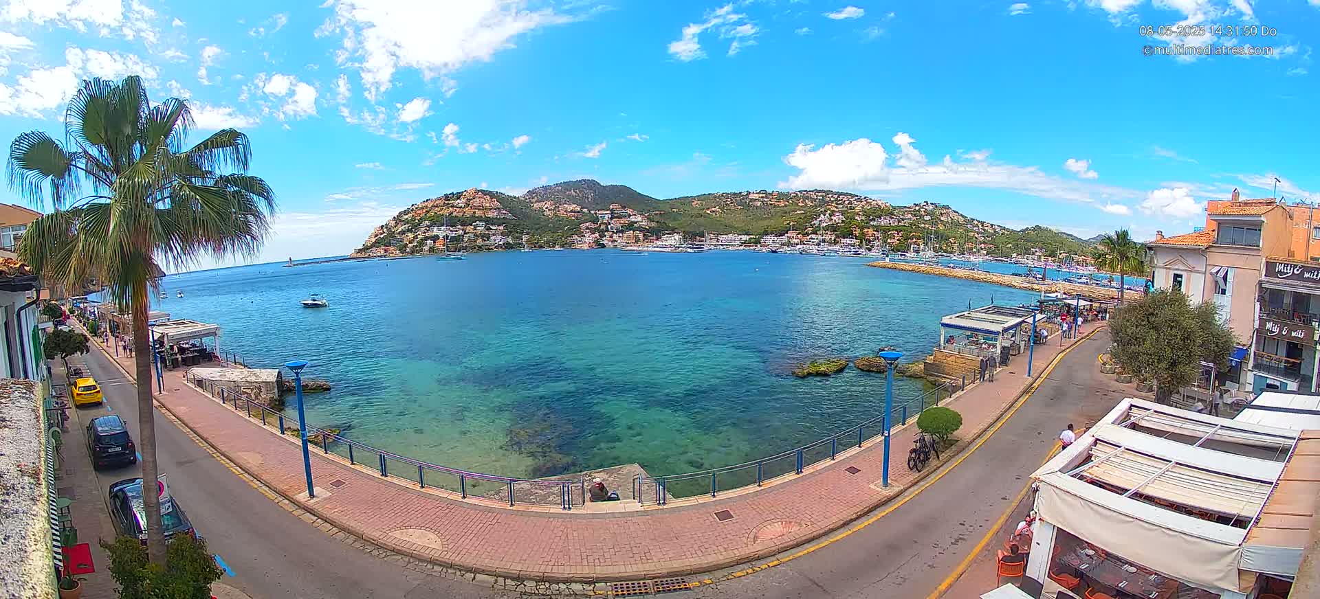 A seaside town under a mostly sunny sky with a calm, clear turquoise bay, boats in the harbor, and palm trees lining a paved walkway.