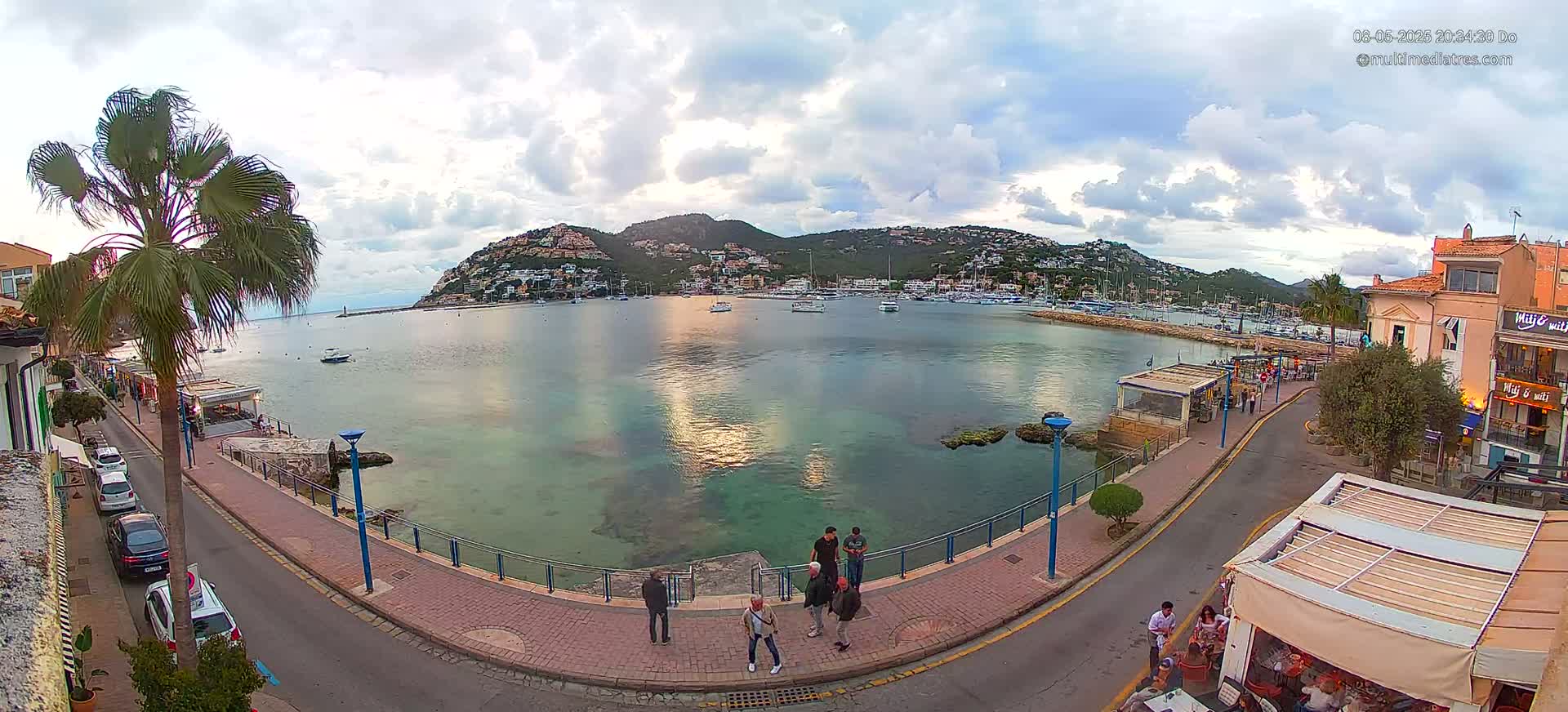 A calm harbor with many boats is nestled below a hillside town under a partly cloudy sky.