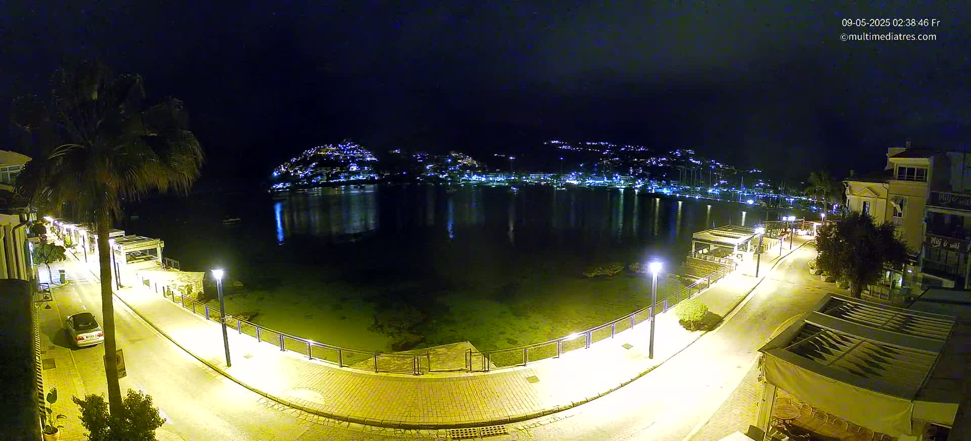 A nighttime, coastal scene shows a calm bay reflecting the lights of a town nestled on a hillside, with a paved walkway and buildings illuminated along the waterfront.